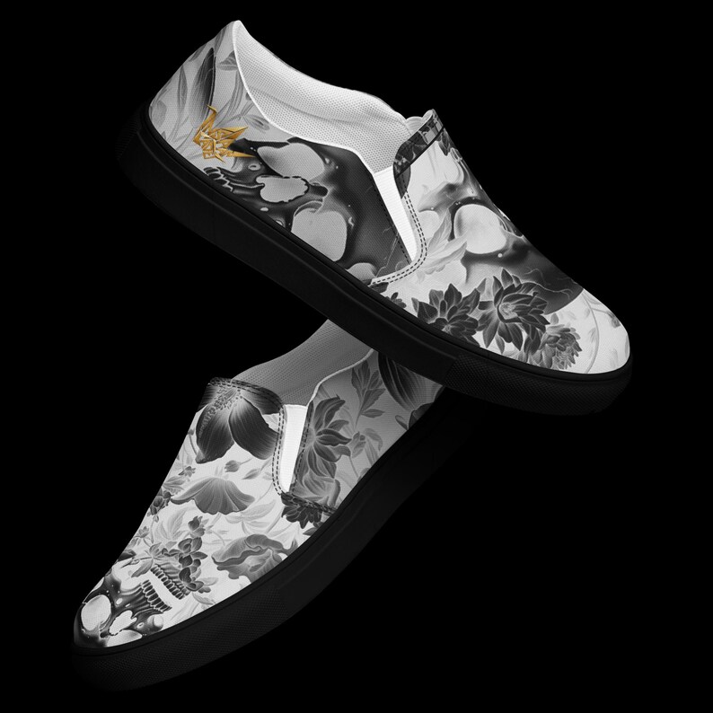 May include: A pair of slip-on sneakers featuring a black and white graphic print of skulls and floral patterns. The shoes have solid black rubber soles and white interior lining. A small gold-colored emblem is visible on the upper side.
