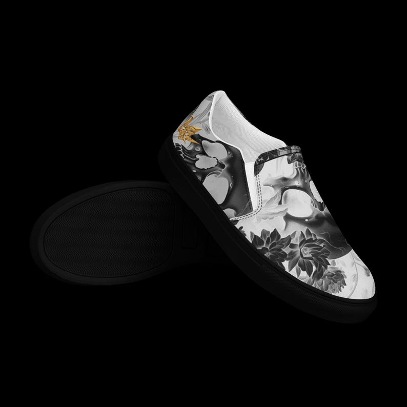 May include: A slip-on shoe featuring a black and white graphic print of a skull surrounded by dark floral elements. The shoe has a solid black rubber sole and a white interior lining. A small gold-colored accent is visible on the side heel area.