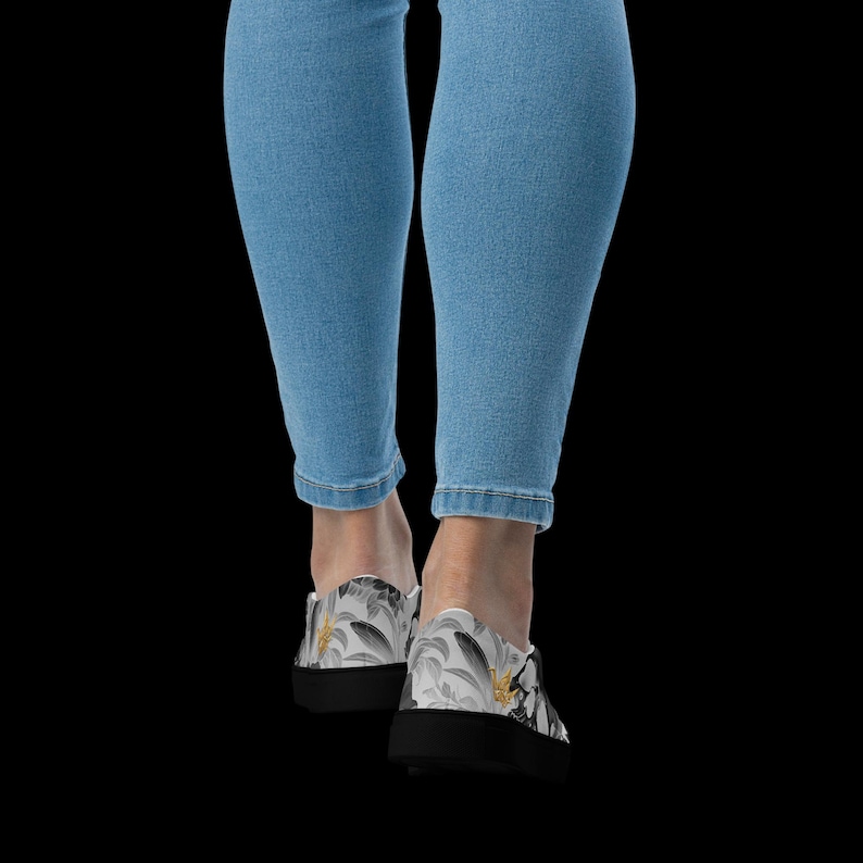 May include: Light blue denim jeans paired with slip-on sneakers featuring a black and white botanical leaf pattern accented with small yellow flowers. The sneakers have a solid black rubber sole and a low-top silhouette with a rounded toe design.