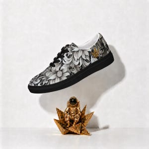 May include: A low-top sneaker featuring a black and white floral pattern across the upper, black laces, and a solid black sole. A small gold emblem is visible on the side. Below the shoe sits a gold-colored astronaut figurine on a star base.