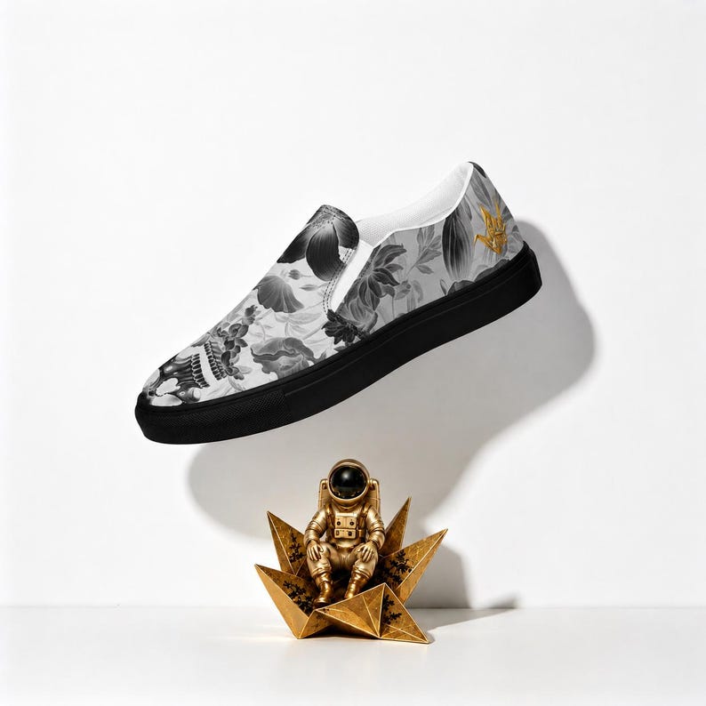 May include: A slip-on sneaker featuring a grayscale floral pattern on the upper, a solid black rubber sole, and a small gold hand gesture icon near the heel. Below the shoe sits a gold-colored astronaut figurine perched on a geometric star base.
