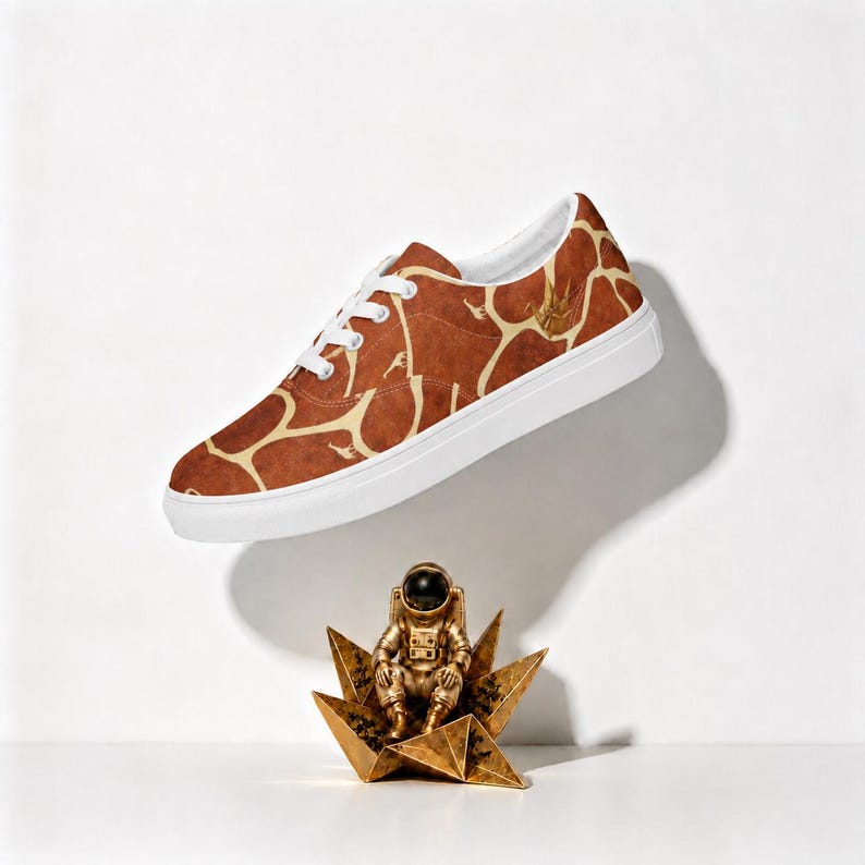 May include: A low-top sneaker featuring a brown and cream giraffe-inspired pattern, white laces, and a thick white rubber sole. Below the shoe sits a small, metallic gold-colored astronaut figurine perched atop a geometric star-shaped base.
