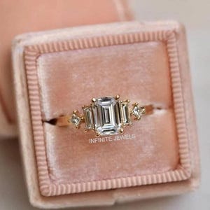 Pode incluir: Um anel de ouro com uma grande pedra central com corte esmeralda e pedras laterais menores. O anel é cravejado em uma banda de ouro e é exibido em uma caixa de anel de veludo rosa. O texto "INFINITE JEWELS" é visível.