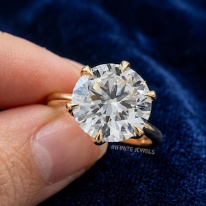 Puede incluir: Un anillo de oro con una gran piedra preciosa transparente de talla brillante redonda, sujeta por seis garras. La banda es de oro amarillo pulido. El texto INFINITE JEWELS es visible en la base del engaste de la piedra preciosa.