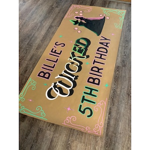 Może przedstawiać: Prostokątny znak z papieru pakowego z napisem "Billie's Wicked 5th Birthday" w kolorze czarnym, białym i zielonym. Znak przedstawia sylwetkę czarownicy i dekoracyjne różowe i zielone akcenty. Znak znajduje się na drewnianej podłodze.