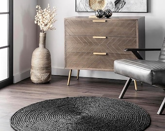 Puede incluir: Una alfombra redonda trenzada de color negro sobre un suelo de madera. El diseño concéntrico de la alfombra es el punto focal. Un aparador de madera con herrajes dorados y una silla de cuero se encuentran en el fondo.