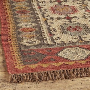 May include: Close-up of a rectangular rug with a detailed pattern. The rug features a color palette of red, brown, beige, and gold. The border has a red hue with a fringe at the bottom. The rug's design includes geometric shapes and patterns.