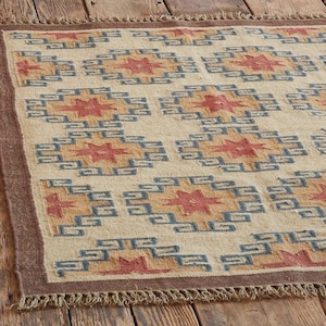 May include: Beige rectangular rug with a geometric pattern in shades of red, blue, and gold. The rug has a brown border and fringe along the edges. The pattern features repeating star-like shapes.