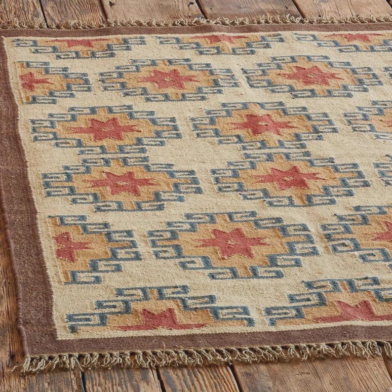 May include: Beige rectangular rug with a geometric pattern in shades of red, blue, and gold. The rug has a brown border and fringe along the edges. The pattern features repeating star-like shapes.