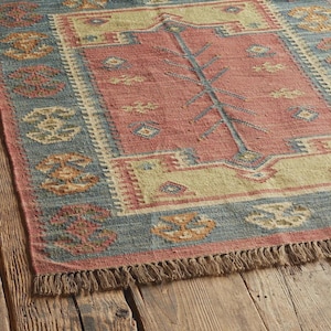 May include: A rectangular area rug with a central red field, bordered by a blue and beige pattern. The rug features a tree-like design in the center and decorative patterns along the edges. The rug has a fringed edge.