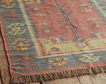 May include: A rectangular area rug with a central red field, bordered by blue and beige geometric patterns. The rug has a fringed edge and is placed on a wooden surface. The design features a tree-like motif.