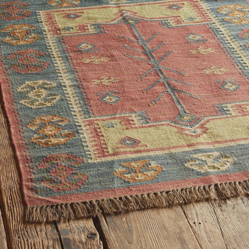 May include: A rectangular area rug with a central red field, bordered by a blue and beige pattern. The rug features a tree-like design in the center and decorative patterns along the edges. The rug has a fringed edge.