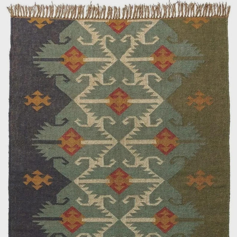 May include: A rectangular rug with a geometric pattern in shades of blue, green, and beige. The design features repeating diamond and arrow shapes with orange accents. The rug has a fringed edge.