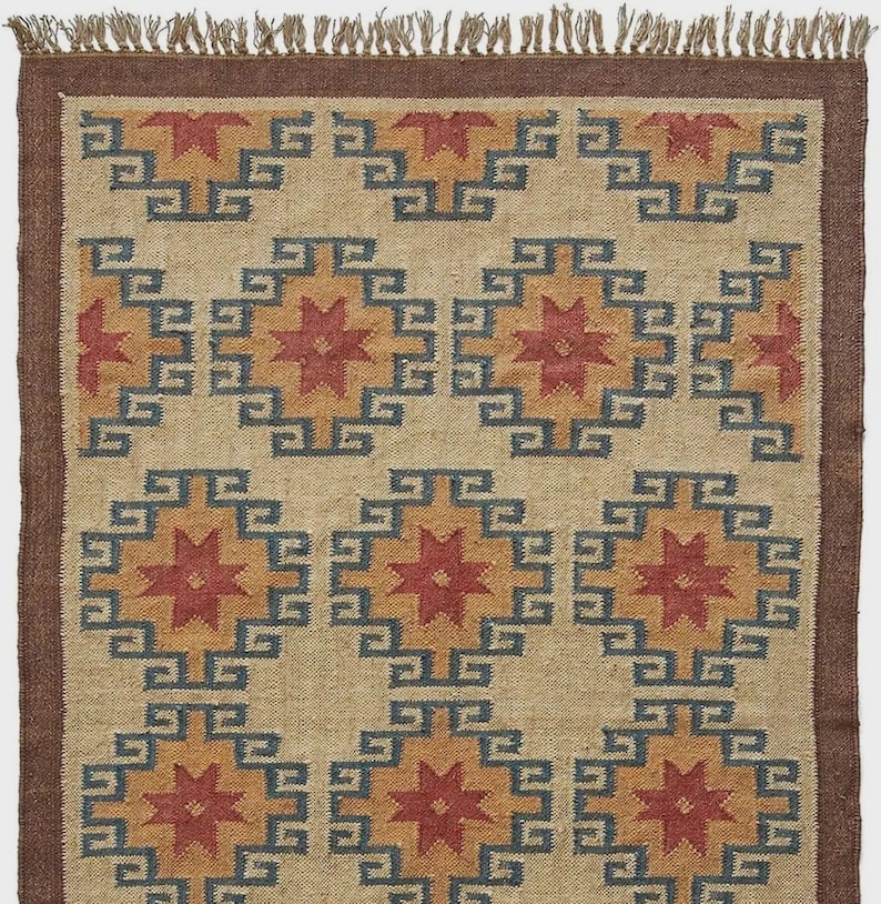 May include: A rectangular jute rug with a geometric pattern. The design features repeating diamond shapes with starburst centers in red and gold, framed by blue squares. The rug has a brown border and fringe.