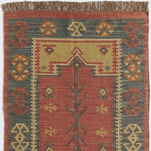 May include: A rectangular rug with a red central field, a green-blue border, and a tan center section. The rug features geometric and floral patterns in shades of tan, orange, and green-blue. The top edge has a brown fringe.