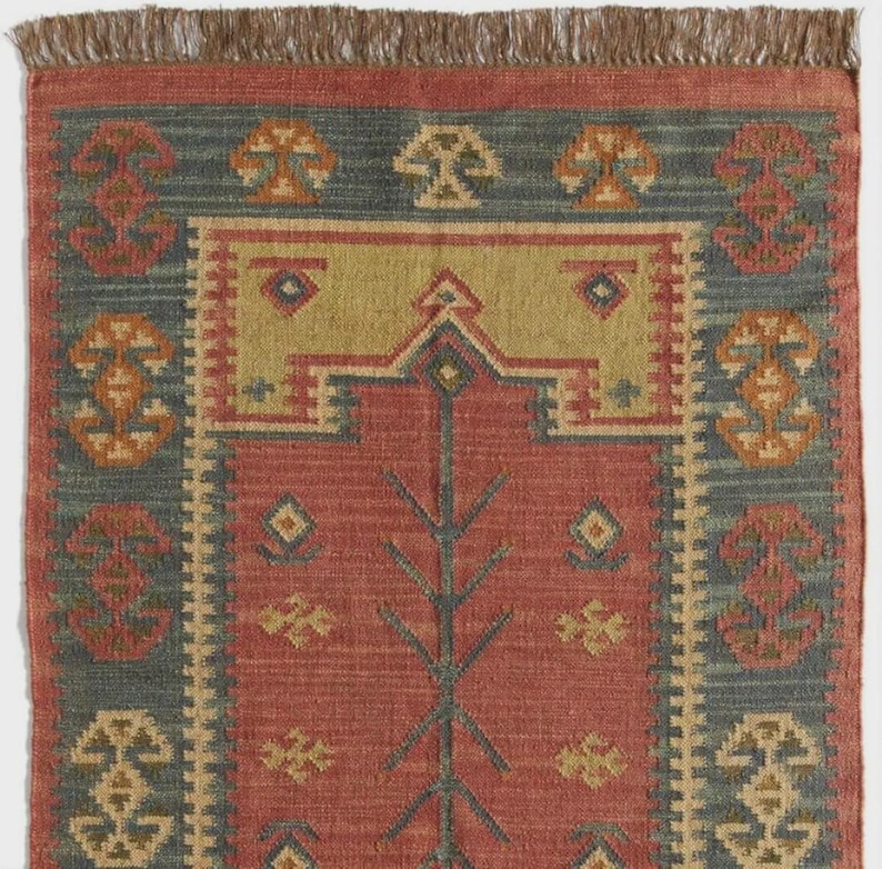May include: A rectangular rug with a red central field, a green-blue border, and a tan center section. The rug features geometric and floral patterns in shades of tan, orange, and green-blue. The top edge has a brown fringe.