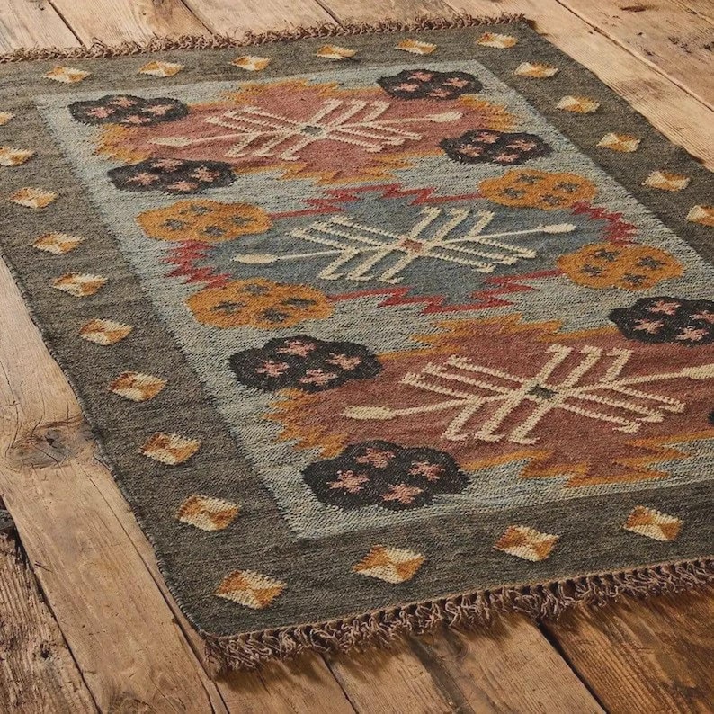 May include: Rectangular area rug with a geometric pattern in shades of gray, blue, orange, and red. The rug has a dark gray border with diamond-shaped accents and fringed edges. The design features stylized floral and geometric shapes.