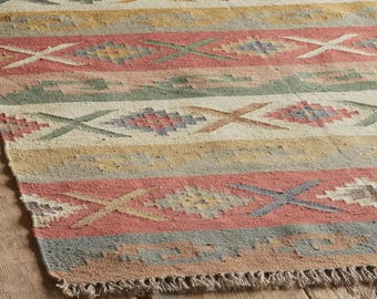 May include: Close-up of a colorful woven rug with a geometric pattern. The rug features horizontal stripes in shades of red, yellow, green, and blue, with an X-shaped design. The rug has a fringed edge and is displayed on a wooden surface.