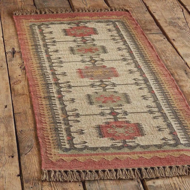 May include: Rectangular area rug with a beige central field and a geometric pattern in red, gray, and brown. The rug has a red border with fringe on the short ends. The rug is made of natural fibers.