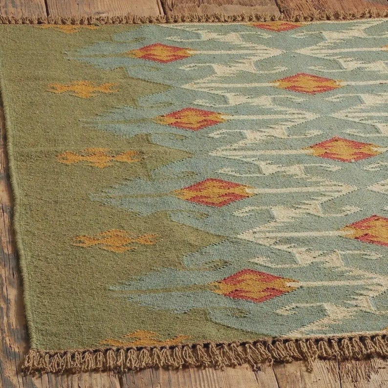May include: A rectangular rug with a geometric pattern. The rug features a green border and a central design in shades of blue, cream, and orange. The rug has a fringed edge and is likely made of woven material.