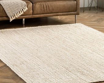 May include: A rectangular, off-white rug with a textured, ribbed design. The rug is made of a natural fiber and is placed on a wooden floor. The rug is a neutral color and would complement various interior design styles.