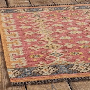 Puede incluir: Una alfombra rectangular con un patrón geométrico en tonos rojos, amarillos y azules. La alfombra tiene un borde con flecos y está hecha de un material tejido. El diseño presenta formas de diamantes y zigzags.