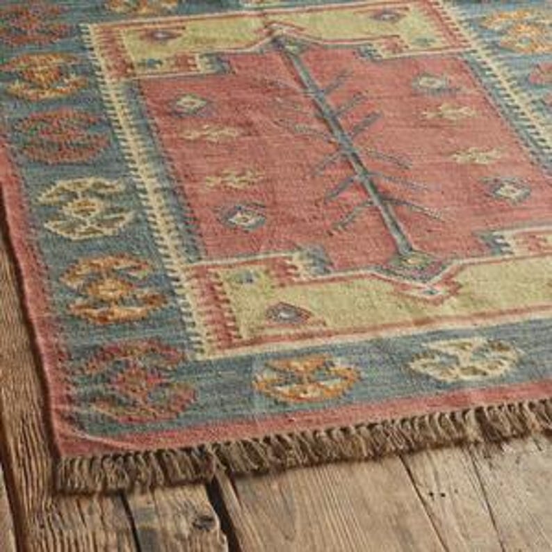 May include: Rectangular rug with a faded red center, bordered by blue and yellow geometric patterns. The rug has a fringed edge and is placed on a wooden surface. The rug's design features a central tree-like motif.