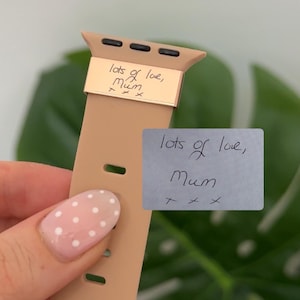 May include: A tan Apple Watch band with a gold-colored metal accent. The metal accent has the handwritten text "lots of love, Mum" engraved on it. A small gray sticky note with the same text is also visible.
