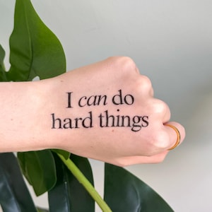 May include: A temporary tattoo on the back of a hand featuring the black text I can do hard things in a serif font. The design is positioned horizontally across the skin, with a thin gold-colored ring visible on the ring finger.