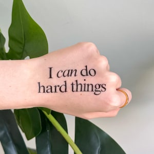 Puede incluir: Un tatuaje temporal en una mano con la frase "I can do hard things" en letras negras. Un anillo dorado adorna el dedo anular. La mano está colocada frente a una gran hoja verde.