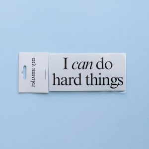 May include: A rectangular white sticker with black text that reads I can do hard things. The sticker is attached to a white backing card with a hanging hole and the vertical text my smile printed in black on the left side of the card.