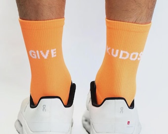 May include: A pair of bright orange ribbed crew socks featuring the white text GIVE on the left sock and KUDOS on the right sock, worn with white athletic sneakers that have black interior lining and a small red logo detail on the heel.