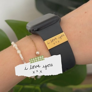 May include: A black smartwatch band with a gold-colored metal accent that reads "i love you" in handwritten script. A white paper with the same text is taped to the wrist. A pearl bracelet and gold chain are also visible.