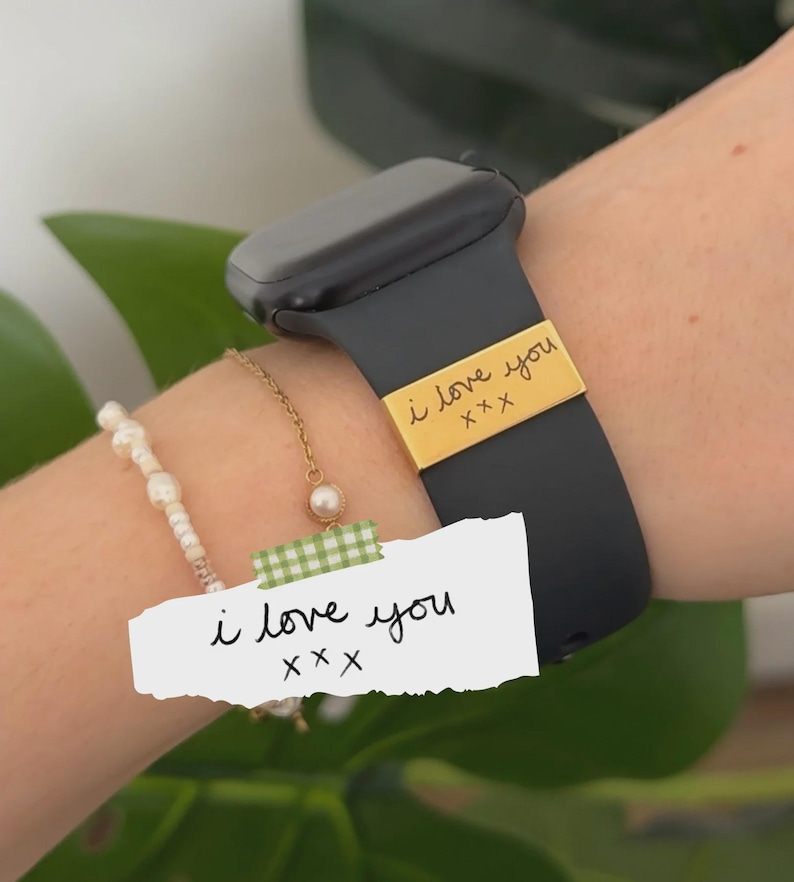 May include: A black smartwatch band with a gold-colored metal accent that reads "i love you" in handwritten script. A white paper with the same text is taped to the wrist. A pearl bracelet and gold chain are also visible.