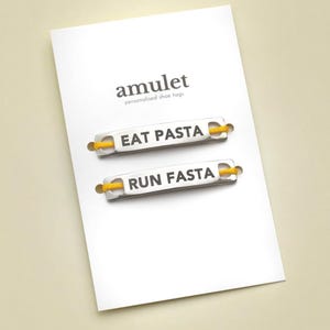 Peut inclure: Une carte blanche avec le mot "amulet" et le texte "personalised shoe tags". Deux étiquettes de chaussures argentées avec des lacets jaunes sont affichées. Une étiquette indique "EAT PASTA" et l'autre "RUN FASTA".