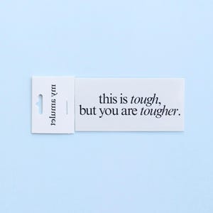 May include: A white rectangular tag with the text "this is tough, but you are tougher" in black. The tag has a small hole for hanging and the words "my summer" printed vertically on the left side. The background is a light blue.