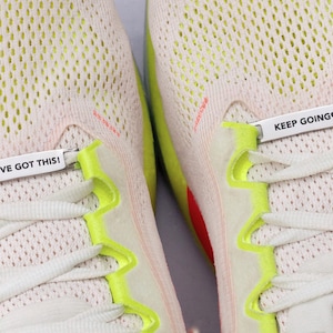 May include: A pair of beige athletic sneakers featuring bright neon yellow lace eyelets and white laces. Two silver metal shoe lace tags are attached to the laces, with the left tag reading YOU'VE GOT THIS! and the right tag reading KEEP GOING&hearts;.