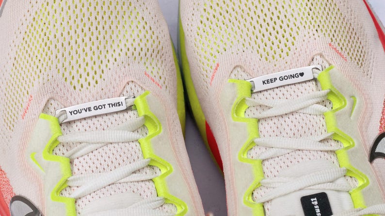 May include: A pair of beige athletic sneakers featuring bright neon yellow lace eyelets and white laces. Two silver metal shoe lace tags are attached to the laces, with the left tag reading YOU'VE GOT THIS! and the right tag reading KEEP GOING&hearts;.