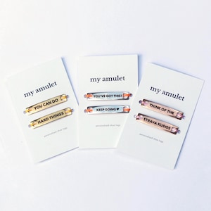 May include: Three white display cards labeled my amulet, each holding two metal shoe tags. The gold tags read YOU CAN DO and HARD THINGS. The silver tags read YOU'VE GOT THIS! and KEEP GOING. The rose gold tags read THINK OF THE and STRAVA KUDOS.