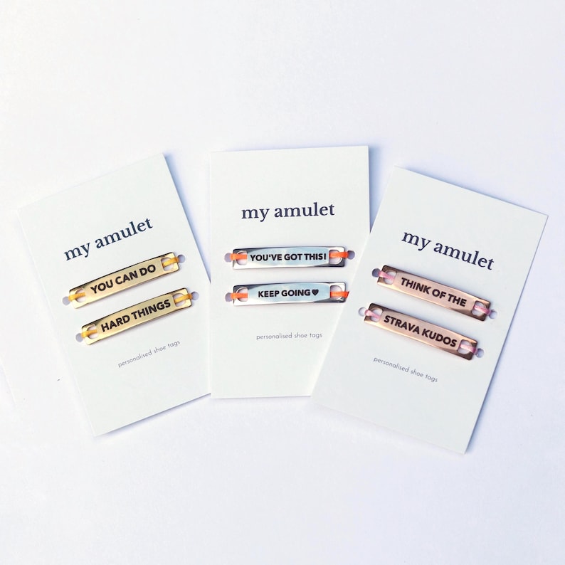 May include: Three white display cards labeled my amulet, each holding two metal shoe tags. The gold tags read YOU CAN DO and HARD THINGS. The silver tags read YOU'VE GOT THIS! and KEEP GOING. The rose gold tags read THINK OF THE and STRAVA KUDOS.