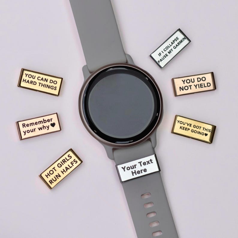 Puede incluir: Un reloj inteligente gris con pantalla negra y correa gris. Se muestran varios dijes rectangulares dorados y plateados con frases motivacionales como "You Can Do Hard Things" y "You've Got This" alrededor del reloj.