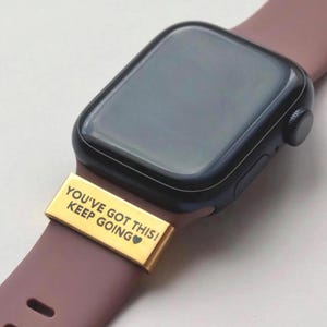 Può includere: Primo piano di uno smartwatch con un cinturino color oro che recita "YOU'VE GOT THIS! KEEP GOING". L'orologio ha uno schermo nero e un cinturino color malva.