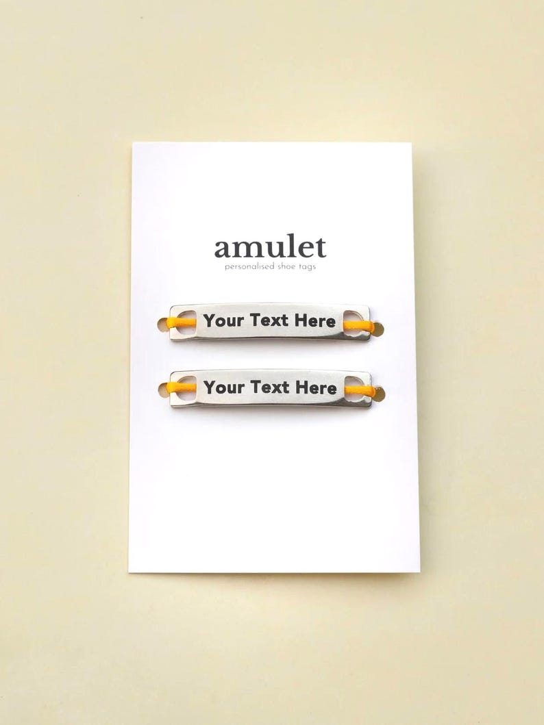 May include: Two silver-colored metal shoe tags with yellow elastic cords, mounted on a white display card. The card features the text amulet personalised shoe tags at the top, and each tag displays the black printed text Your Text Here.