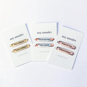 Op de afbeelding: Drie witte presentatiekaarten met het opschrift my amulet, elk met twee rechthoekige metalen schoenlabels. De labels bevatten gegraveerde teksten zoals YOU CAN DO, HARD THINGS, YOU'VE GOT THIS!, KEEP GOING, THINK OF THE en STRAVA KUDOS in goud, zilver en roségoud.
