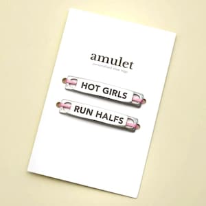 May include: A white card featuring the word "amulet" and "personalised shoe tags" in black. Two silver shoe tags with pink laces are visible, with the text "HOT GIRLS" and "RUN HALFS".