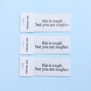May include: Three white rectangular cards with the text "this is tough, but you are tougher" printed in black. Each card has a small, attached section with a hole for hanging. The background is a light blue.