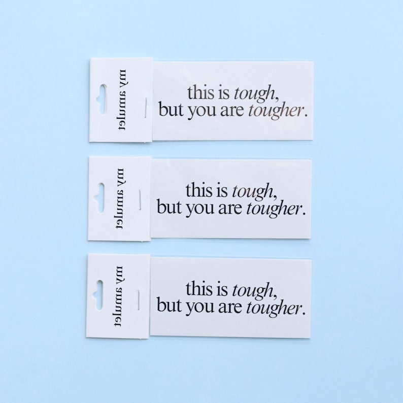 May include: Three white rectangular cards with the text "this is tough, but you are tougher" printed in black. Each card has a small, attached section with a hole for hanging. The background is a light blue.
