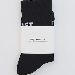 May include: Black socks with white text reading "LAST" are wrapped with a white band. The band features the text "my amulet" and the phrase "a beacon of strength, resilience and determination."