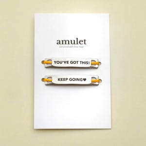 Op de afbeelding: Een witte kaart met het woord "amulet" en de tekst "personalised shoe tags". Er worden twee zilveren schoenlabels met gele veters weergegeven. Op één label staat "YOU'VE GOT THIS!" en op de andere "KEEP GOING ♥".