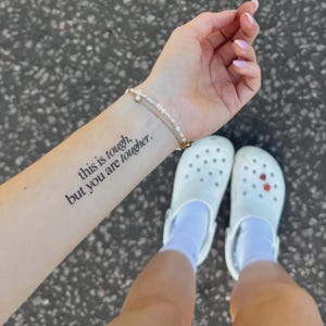 May include: A person's arm with a small tattoo that reads "this is tough, but you are tougher." A delicate bracelet adorns the wrist. The person is wearing white clogs with red marks and white socks. The background is a textured gray surface.